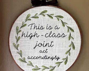 May include: Embroidered hoop art with a white background and green leaves surrounding the text "This is a high-class joint act accordingly".