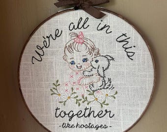 May include: A circular embroidery hoop featuring a beige fabric background with a brown ribbon bow at the top. The design shows a child holding a small dog, surrounded by daisies, with the text "We're all in this together -like hostages-".