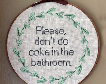 May include: A white embroidery hoop with a green leafy wreath around the edge. The text inside the wreath reads "Please don't do coke in the bathroom."