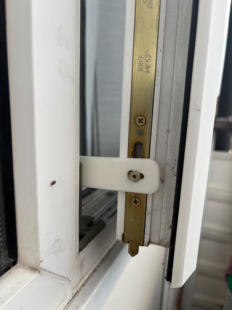 Op de afbeelding: Close-up van een wit raamkozijn met een messingkleurige metalen grendel en een witte plastic handgreep. De grendel is met schroeven bevestigd. Het frame vertoont tekenen van slijtage.