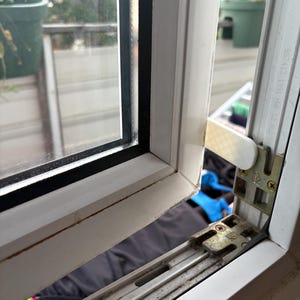 Op de afbeelding: Een close-up van een wit raamkozijn met een zwarte rand rond het glas. Het raam staat gedeeltelijk open en toont het mechanisme en een glimp van de buitenomgeving, inclusief een groene plant in een pot.