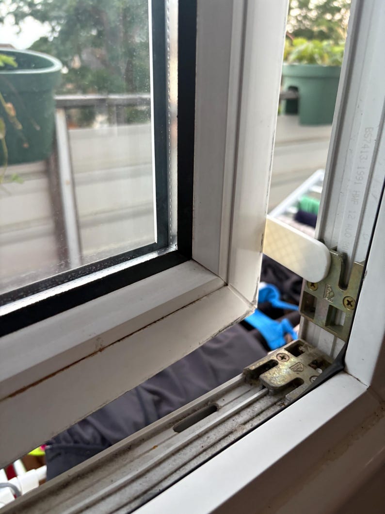 Op de afbeelding: Een close-up van een wit raamkozijn met een zwarte rand rond het glas. Het raam staat gedeeltelijk open en toont het mechanisme en een glimp van de buitenomgeving, inclusief een groene plant in een pot.