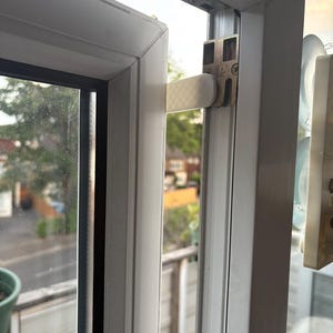 Op de afbeelding: Close-up van een wit raamkozijn met een gedeeltelijk open raam. Het raam heeft een metalen scharnier en een witte hendel. De achtergrond toont een straat en bomen.