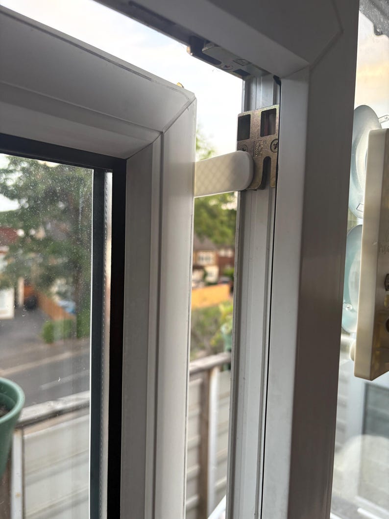 Op de afbeelding: Close-up van een wit raamkozijn met een gedeeltelijk open raam. Het raam heeft een metalen scharnier en een witte hendel. De achtergrond toont een straat en bomen.