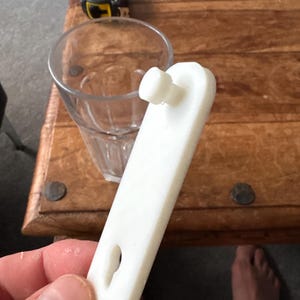 Op de afbeelding: Een wit plastic object met een pin en een sleutelgatvormige opening. Een helder glas en een geel en zwart meetlint staan op de achtergrond. Het object wordt in een hand gehouden.