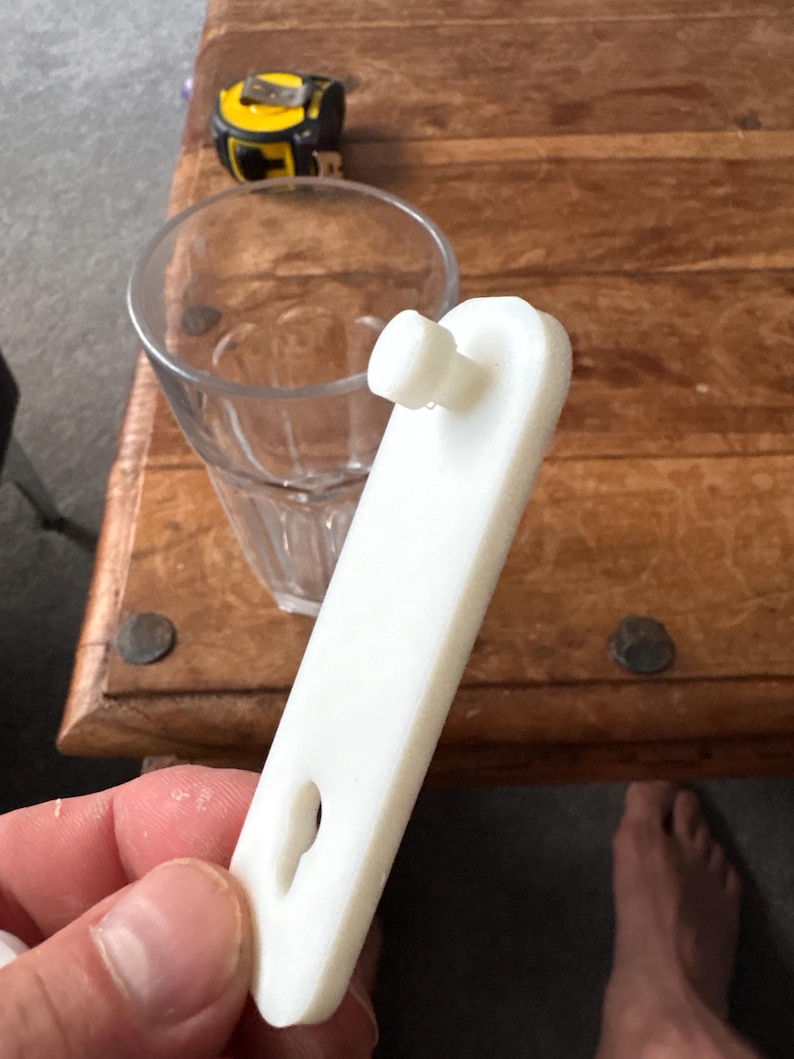 Op de afbeelding: Een wit plastic object met een pin en een sleutelgatvormige opening. Een helder glas en een geel en zwart meetlint staan op de achtergrond. Het object wordt in een hand gehouden.