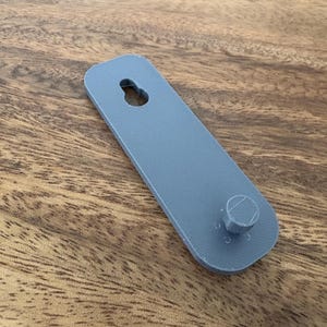 Op de afbeelding: Een grijs, rechthoekig object met een gat aan de bovenkant en een cilindrische uitsteeksel aan de onderkant. Het object is gemaakt van een stevig materiaal, waarschijnlijk een montagebeugel. Het ligt op een houten oppervlak.