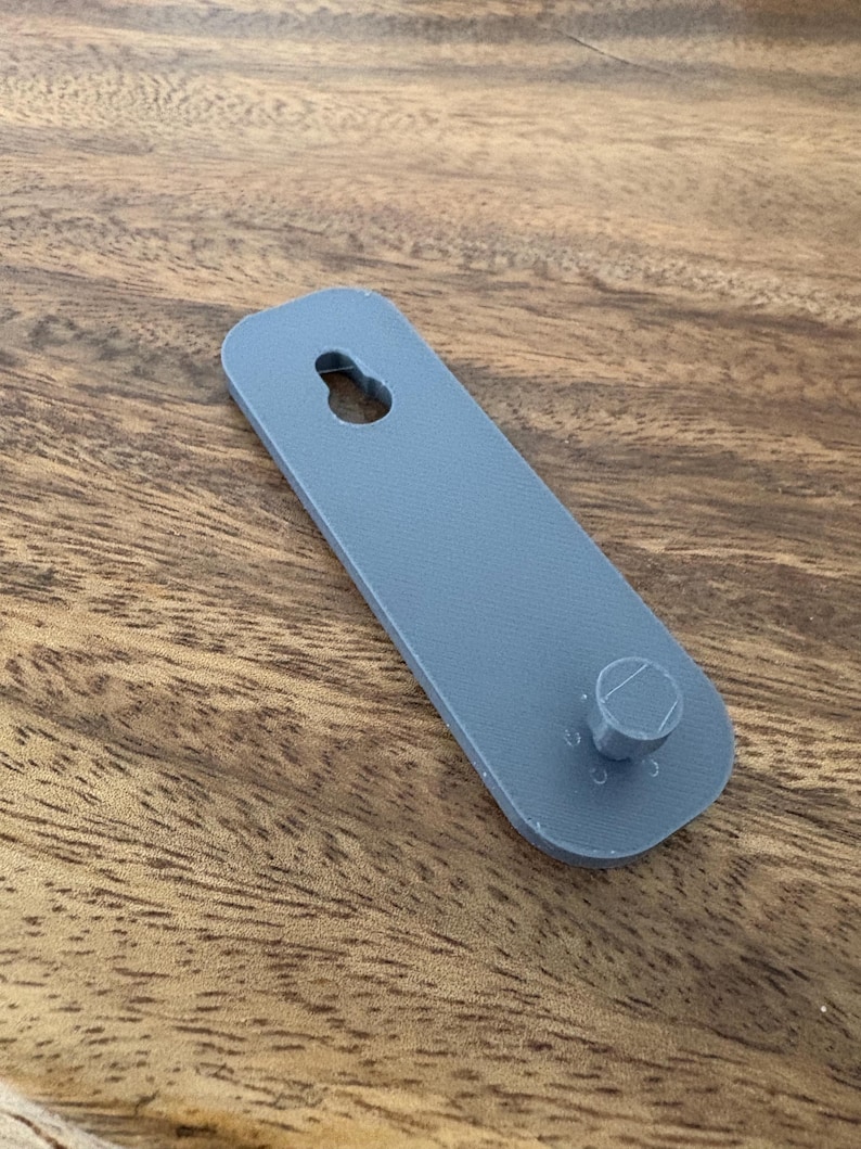 Op de afbeelding: Een grijs, rechthoekig object met een gat aan de bovenkant en een cilindrische uitsteeksel aan de onderkant. Het object is gemaakt van een stevig materiaal, waarschijnlijk een montagebeugel. Het ligt op een houten oppervlak.