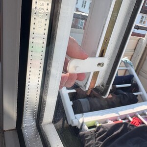 Op de afbeelding: Een close-up van een wit raamkozijn met een witte plastic grendel. Het raam is gedeeltelijk open en onthult een uitzicht op daken en een wasrek. Het woord "GLASS" is zichtbaar op het frame.