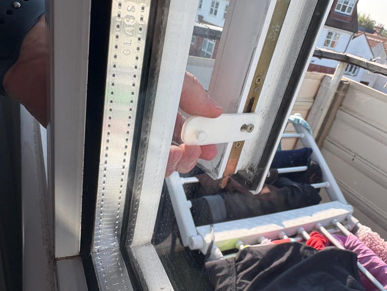 Op de afbeelding: Een close-up van een wit raamkozijn met een witte plastic grendel. Het raam is gedeeltelijk open en onthult een uitzicht op daken en een wasrek. Het woord "GLASS" is zichtbaar op het frame.