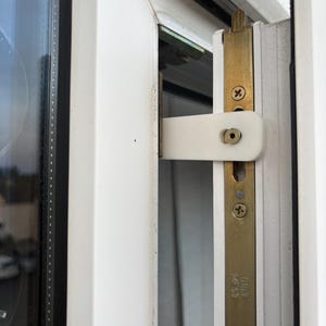 Op de afbeelding: Close-up van een witte raamgrendel en een messingkleurige metalen plaat met schroeven. Het raamkozijn is wit en het glas is zichtbaar aan de linkerkant. De grendel staat dicht.