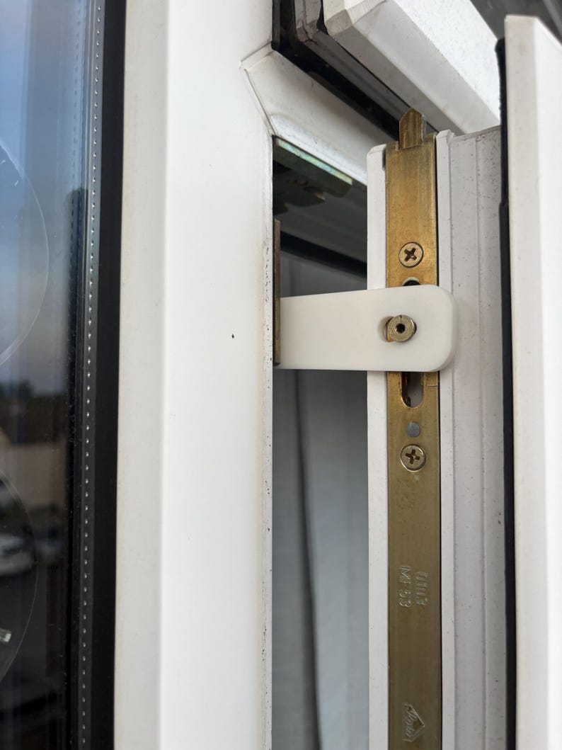 Op de afbeelding: Close-up van een witte raamgrendel en een messingkleurige metalen plaat met schroeven. Het raamkozijn is wit en het glas is zichtbaar aan de linkerkant. De grendel staat dicht.
