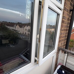 Op de afbeelding: Een wit dubbelglasraam staat gedeeltelijk open en onthult een uitzicht op huizen en een bewolkte lucht. Een doorzichtig zuignapapparaat is aan het glas bevestigd. Het raamkozijn is wit en de buitenmuur is van baksteen.