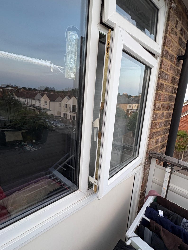 Op de afbeelding: Een wit dubbelglasraam staat gedeeltelijk open en onthult een uitzicht op huizen en een bewolkte lucht. Een doorzichtig zuignapapparaat is aan het glas bevestigd. Het raamkozijn is wit en de buitenmuur is van baksteen.