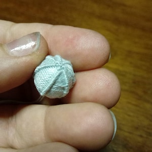 Può includere: Una piccola palla di filo blu chiaro, tenuta tra le dita. Il filo è strettamente avvolto, creando una superficie strutturata. La palla misura circa 1,3 cm di diametro, con dettagli del filo visibili. Lo sfondo è una superficie di legno sfocata.