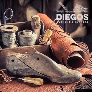 Może przedstawiać: Rustykalna scena warsztatowa z narzędziami szewskimi. Drewniany kopyto szewskie, szpule nici, pędzle i rolka sk&oacute;ry ułożone na drewnianej powierzchni. Obraz zawiera tekst "DIEGOS AUTHENTIC AVARCAS".