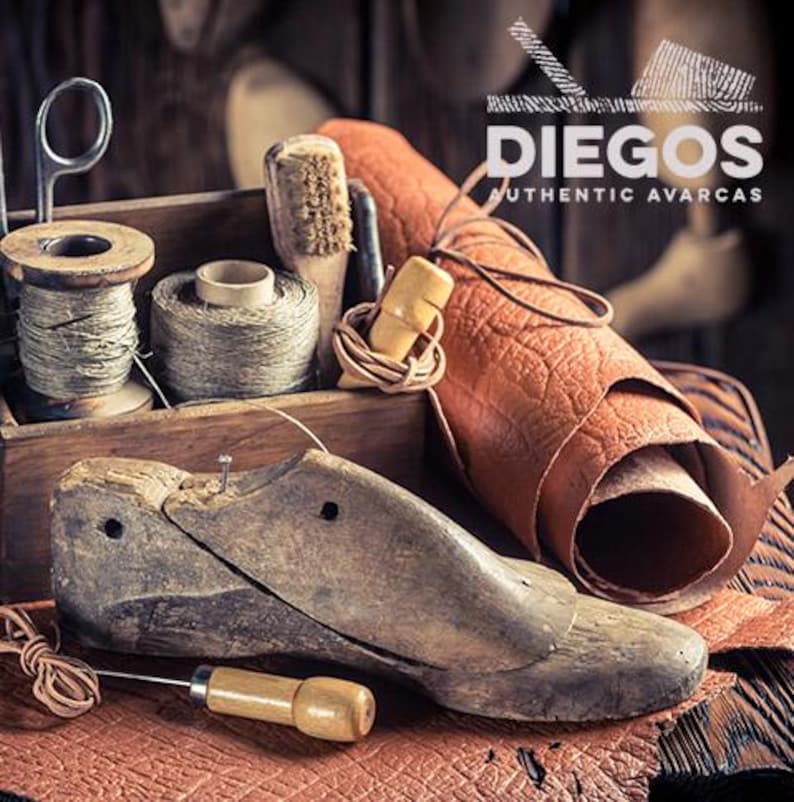 Może przedstawiać: Rustykalna scena warsztatowa z narzędziami szewskimi. Drewniany kopyto szewskie, szpule nici, pędzle i rolka sk&oacute;ry ułożone na drewnianej powierzchni. Obraz zawiera tekst "DIEGOS AUTHENTIC AVARCAS".