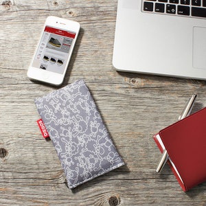 May include: A gray phone case with a white sheep pattern. The case has a red label with the word "DIEGOS" on it. The case is lying on a wooden surface next to a red notebook and a silver pen.