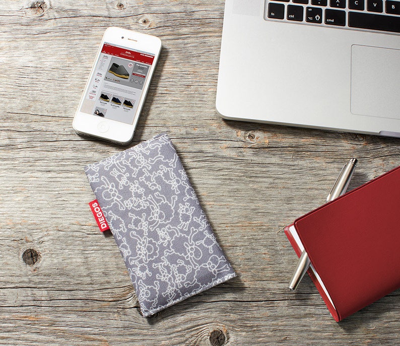 May include: A gray phone case with a white sheep pattern. The case has a red label with the word "DIEGOS" on it. The case is lying on a wooden surface next to a red notebook and a silver pen.