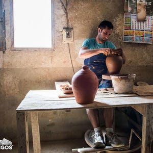 May include: A potter working on a clay vase in a rustic workshop. The artisan is wearing a blue apron and shaping the vase on a pottery wheel. A finished terracotta vase sits on a wooden table, along with other pottery items.