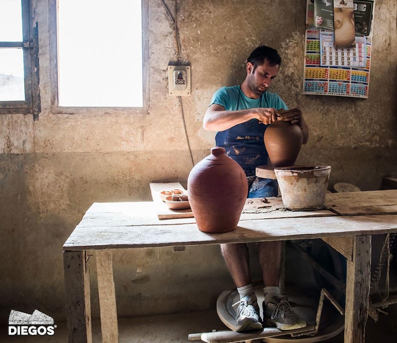 May include: A potter working on a clay vase in a rustic workshop. The artisan is wearing a blue apron and shaping the vase on a pottery wheel. A finished terracotta vase sits on a wooden table, along with other pottery items.