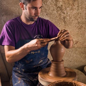 May include: A potter in a purple shirt and blue apron shapes a brown clay vase on a spinning pottery wheel. The image includes the text "DIEGOS" in the upper right corner. A bowl of wet clay sits in the foreground.