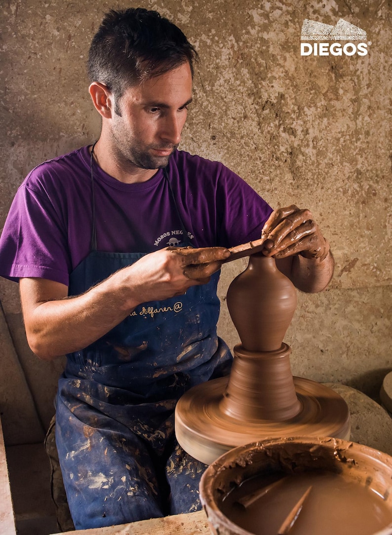 May include: A potter in a purple shirt and blue apron shapes a brown clay vase on a spinning pottery wheel. The image includes the text "DIEGOS" in the upper right corner. A bowl of wet clay sits in the foreground.