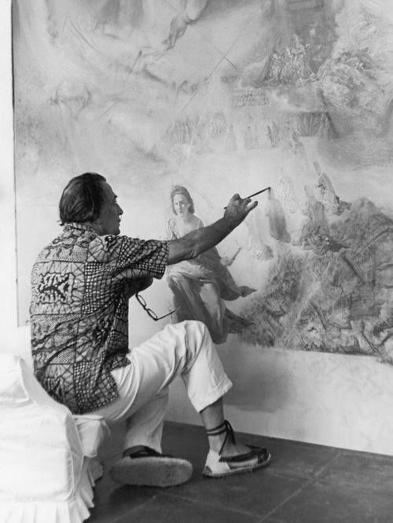May include: A man in a patterned shirt and white pants is painting a large mural on a wall. He is using a brush and is looking at the mural. The mural is a black and white image of a scene with many figures.