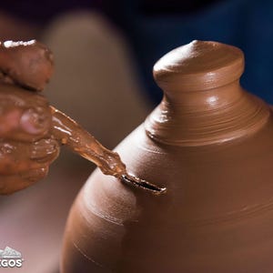 May include: A potter's hands shaping a brown clay pot on a pottery wheel. The potter uses a tool to refine the shape of the pot, creating a smooth, curved surface. The image captures the process of pottery making.