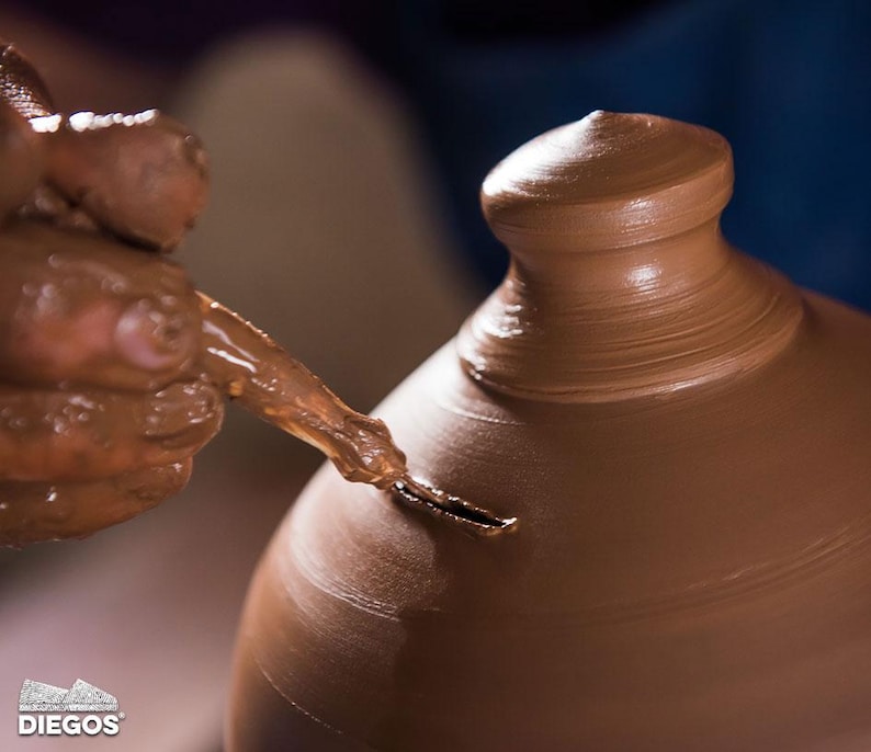 May include: A potter's hands shaping a brown clay pot on a pottery wheel. The potter uses a tool to refine the shape of the pot, creating a smooth, curved surface. The image captures the process of pottery making.