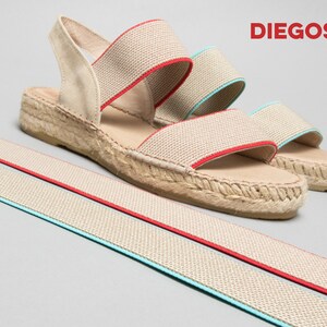 May include: Beige espadrille sandals with red and blue elastic straps. The sandals have a jute wedge sole and are made by the brand "DIEGOS".