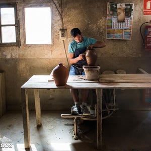 May include: A person working on a pottery wheel in a workshop. The person is wearing a blue shirt and apron, shaping a brown clay pot. A finished pot sits on the table. The workshop has a rustic, aged appearance with natural light.