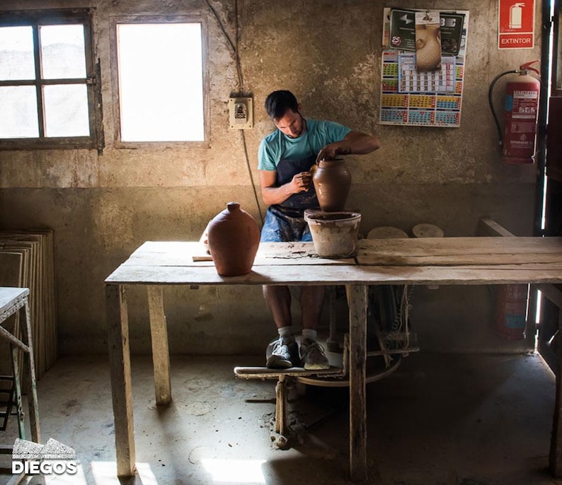 May include: A person working on a pottery wheel in a workshop. The person is wearing a blue shirt and apron, shaping a brown clay pot. A finished pot sits on the table. The workshop has a rustic, aged appearance with natural light.