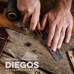 Puede incluir: Primer plano de las manos de una persona trabajando en un proyecto de cuero. Est&aacute;n usando un peque&ntilde;o cuchillo para cortar el cuero y una regla para medirlo. El cuero es marr&oacute;n y las herramientas son plateadas. El fondo es una mesa de madera con un carrete de hilo y otras herramientas.