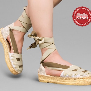 May include: Beige espadrille sandals with olive green straps and a woven jute sole. The sandals are tied around the ankle with a ribbon. The brand name "DIEGOS" is on a red circle label with the text "AUTHENTIC ESPADRILLES HANDMADE IN SPAIN".