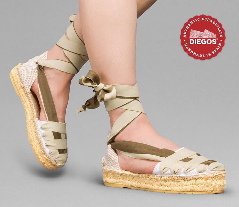 May include: Beige espadrille sandals with olive green straps and a woven jute sole. The sandals are tied around the ankle with a ribbon. The brand name "DIEGOS" is on a red circle label with the text "AUTHENTIC ESPADRILLES HANDMADE IN SPAIN".