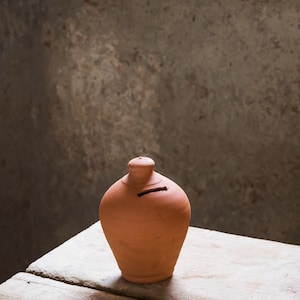 May include: A terracotta piggy bank with a slot for coins. The bank has a rounded body and a small, cylindrical neck. The bank is sitting on a weathered wooden surface. The logo "DIEGOS" is in the upper right corner.