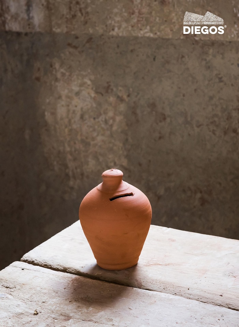 May include: A terracotta piggy bank with a slot for coins. The bank has a rounded body and a small, cylindrical neck. The bank is sitting on a weathered wooden surface. The logo "DIEGOS" is in the upper right corner.