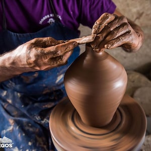 May include: A potter shapes a brown clay vase on a spinning wheel. The potter's hands are covered in wet clay as they use a tool to refine the vase's form. The image captures the process of pottery making, highlighting the craft and skill involved.