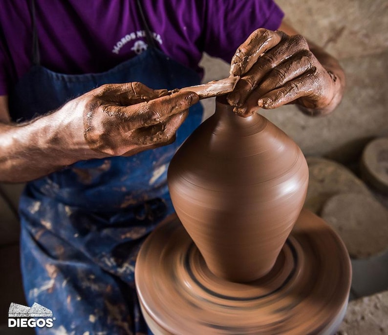 May include: A potter shapes a brown clay vase on a spinning wheel. The potter's hands are covered in wet clay as they use a tool to refine the vase's form. The image captures the process of pottery making, highlighting the craft and skill involved.