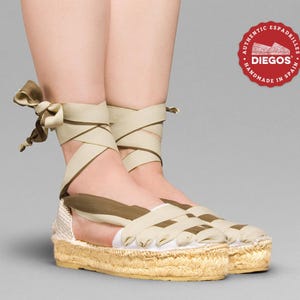 May include: A pair of beige espadrille sandals with brown straps. The straps are tied around the ankles and cross over the top of the foot. The sandals have a jute platform sole. The sandals are handmade in Spain.