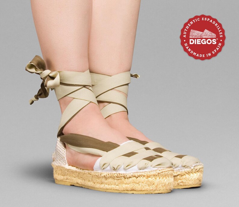 May include: A pair of beige espadrille sandals with brown straps. The straps are tied around the ankles and cross over the top of the foot. The sandals have a jute platform sole. The sandals are handmade in Spain.