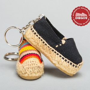 Peut inclure: Un porte-clés en forme de paire d'espadrilles miniatures. Une chaussure est noire avec une semelle en jute et l'autre est multicolore avec une semelle en jute. Les porte-clés sont attachés à un anneau de clé argenté. Les porte-clés présentent le logo "DIEGOS" et le texte "AUTHENTIC ESPADRILLES HANDMADE IN SPAIN".