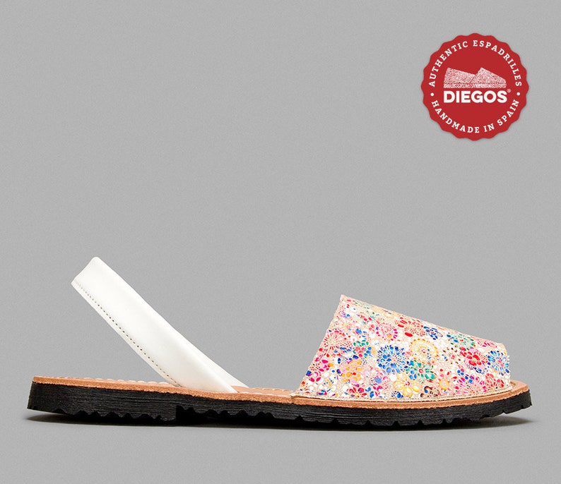 Może przedstawiać: Pojedynczy sandał espadryle z kwiatowym wzorem w kolorze r&oacute;żowym, niebieskim, ż&oacute;łtym i białym. Sandał ma biały pasek, brązową wkładkę i czarną podeszwę. Logo marki jest widoczne w prawym g&oacute;rnym rogu.