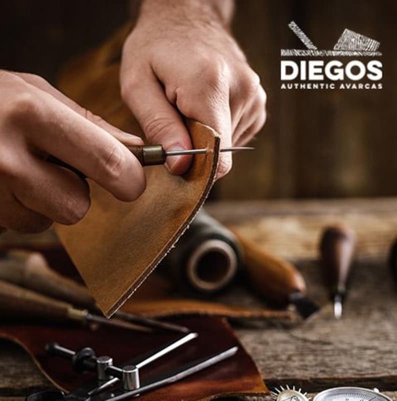 Puede incluir: Primer plano de las manos de una persona trabajando con cuero usando una herramienta para perforar agujeros en el material. El cuero es marr&oacute;n y la herramienta est&aacute; hecha de madera y metal. El fondo es una mesa de madera con otras herramientas de trabajo del cuero.