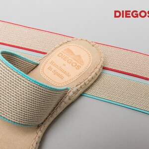 May include: Beige espadrille sandals with a blue elastic strap and a brown leather label that reads "DIEGOS Handmade in Spain".