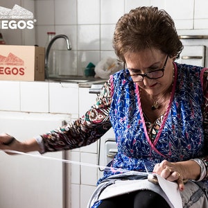 May include: A woman in a blue and pink patterned dress sits in a kitchen and sews a piece of fabric. She is using a white thread and a needle. The fabric is white and has a blue pattern. There is a box with the logo "DIEGOS" on it in the background.
