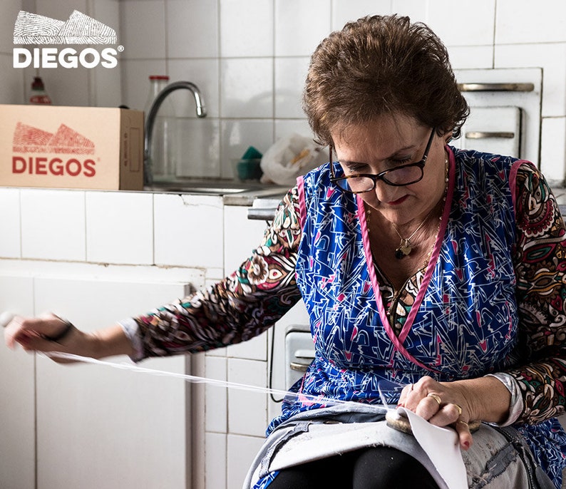 May include: A woman in a blue and pink patterned dress sits in a kitchen and sews a piece of fabric. She is using a white thread and a needle. The fabric is white and has a blue pattern. There is a box with the logo "DIEGOS" on it in the background.