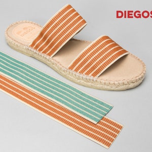 Puede incluir: Un par de sandalias de esparto marrones y beige con dos tiras de rayas naranjas y blancas. Las sandalias est&aacute;n sobre un fondo gris claro. El nombre de la marca "DIEGOS" est&aacute; impreso en rojo en la esquina superior derecha de la imagen.