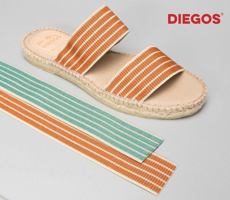 Puede incluir: Un par de sandalias de esparto marrones y beige con dos tiras de rayas naranjas y blancas. Las sandalias est&aacute;n sobre un fondo gris claro. El nombre de la marca "DIEGOS" est&aacute; impreso en rojo en la esquina superior derecha de la imagen.